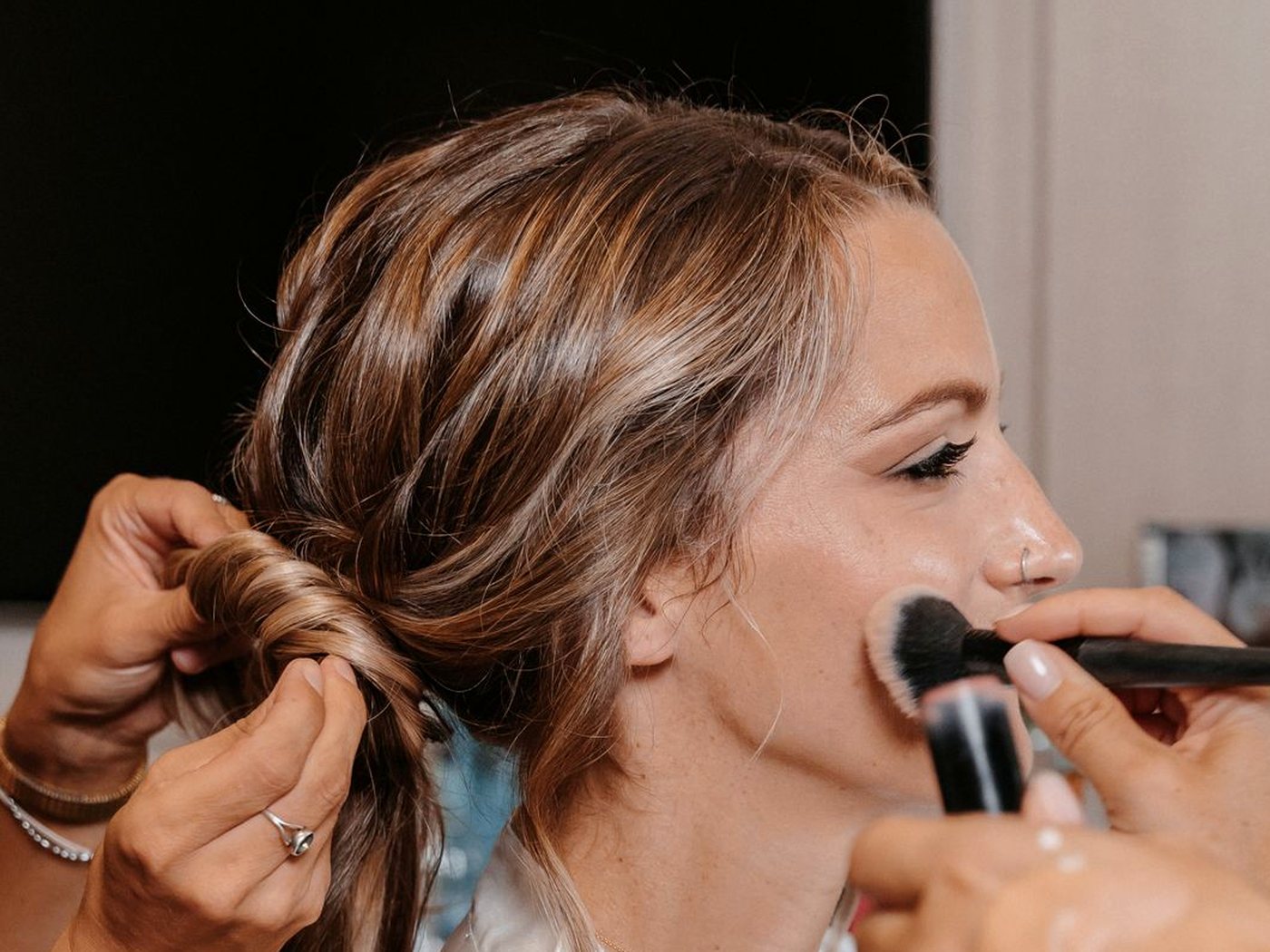 Une coiffeuse noue un chignon pendant qu’un pinceau maquille la joue de la cliente