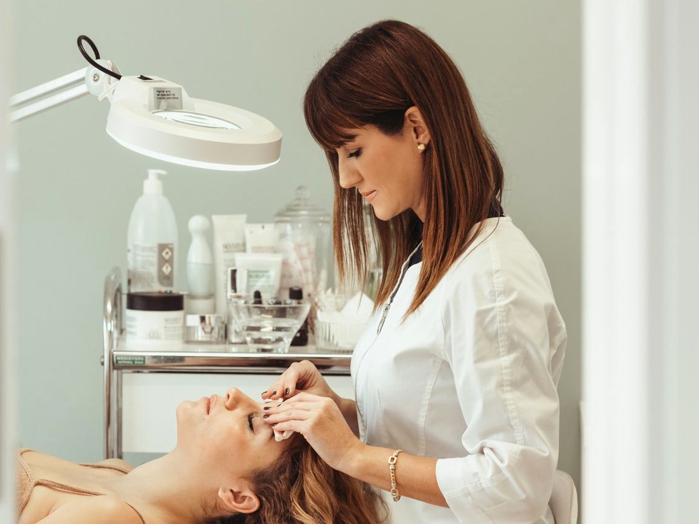 Une esthéticienne en blouse blanche nettoie le visage d’une cliente allongée, sous une lampe loupe