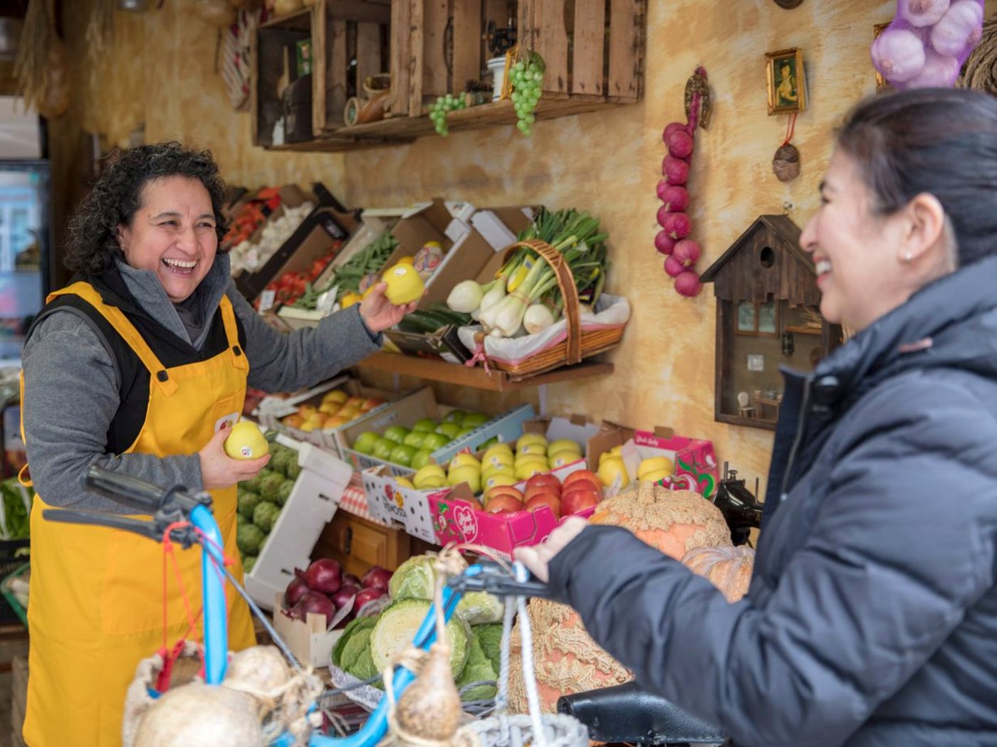 Une marchande de fruits et légumes en tablier jaune rit avec une cliente devant son étal