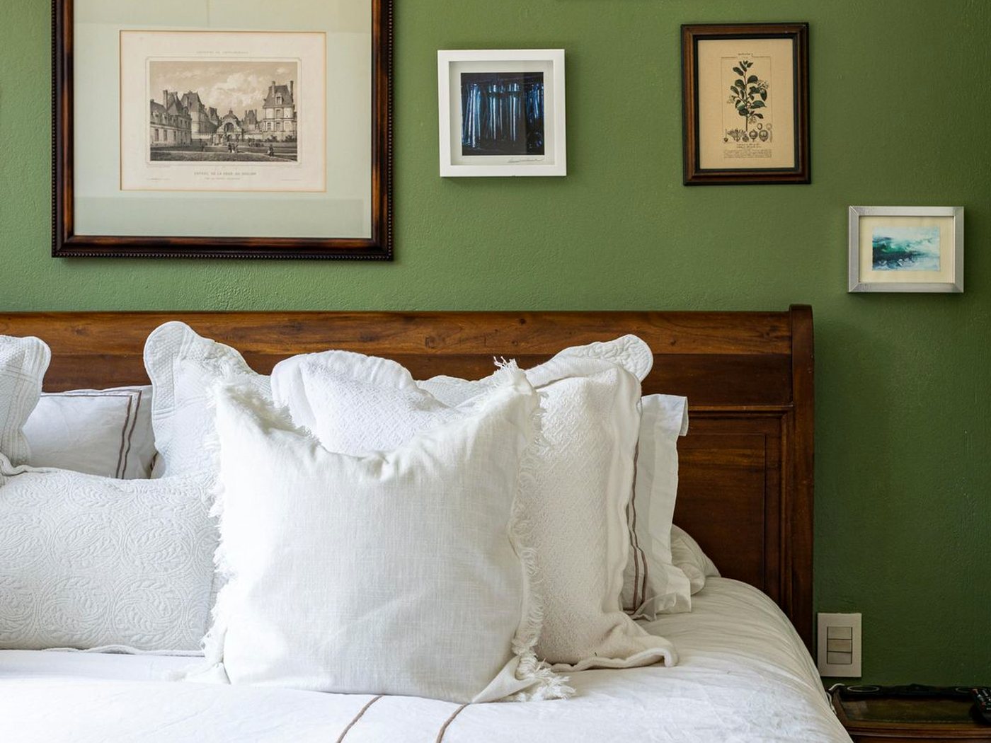 Un lit aux oreillers blancs sous des gravures encadrées, contre un mur vert